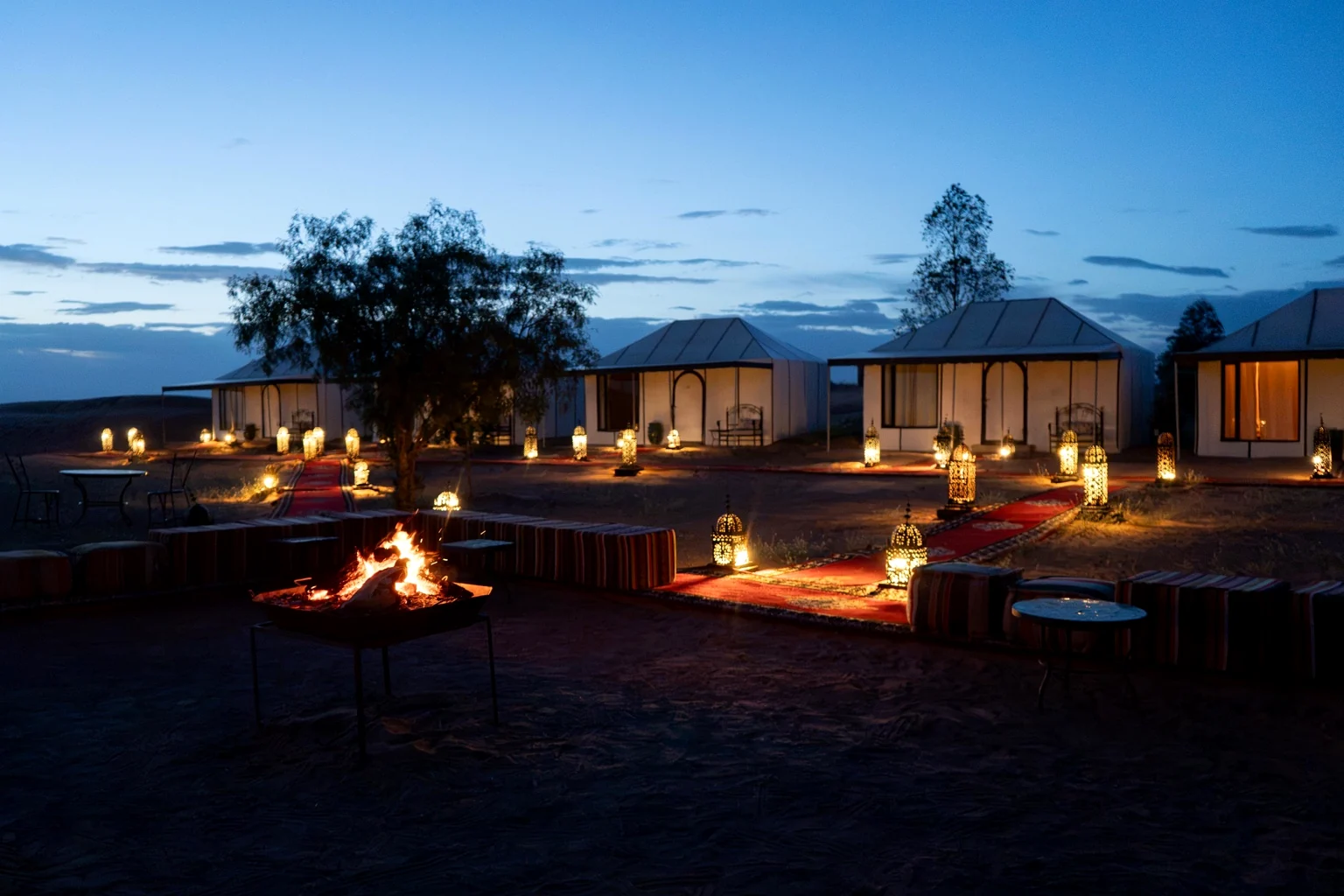 Campfire burning beside low cushioned seating with lit tents behind at blue hour in the Merzouga desert