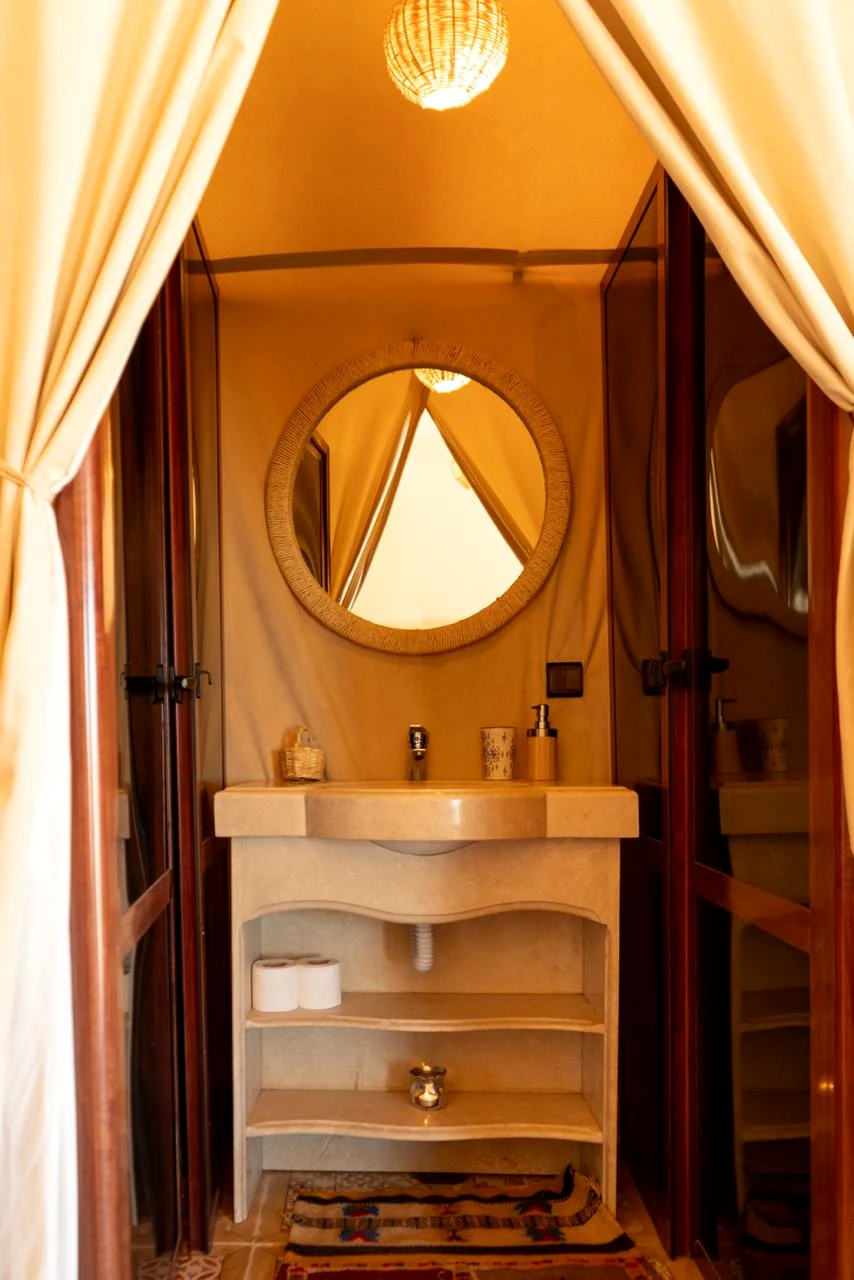 Private ensuite tent bathroom with stone washbasin, round rattan mirror and warm lantern light